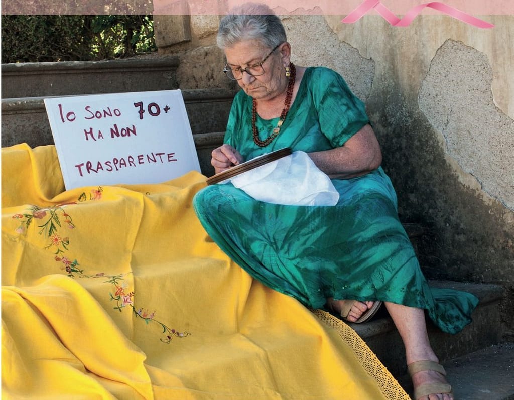 Una donna over 70 ricama seduta sui gradini accanto al cartello "Io sono 70+ ma non trasparente"